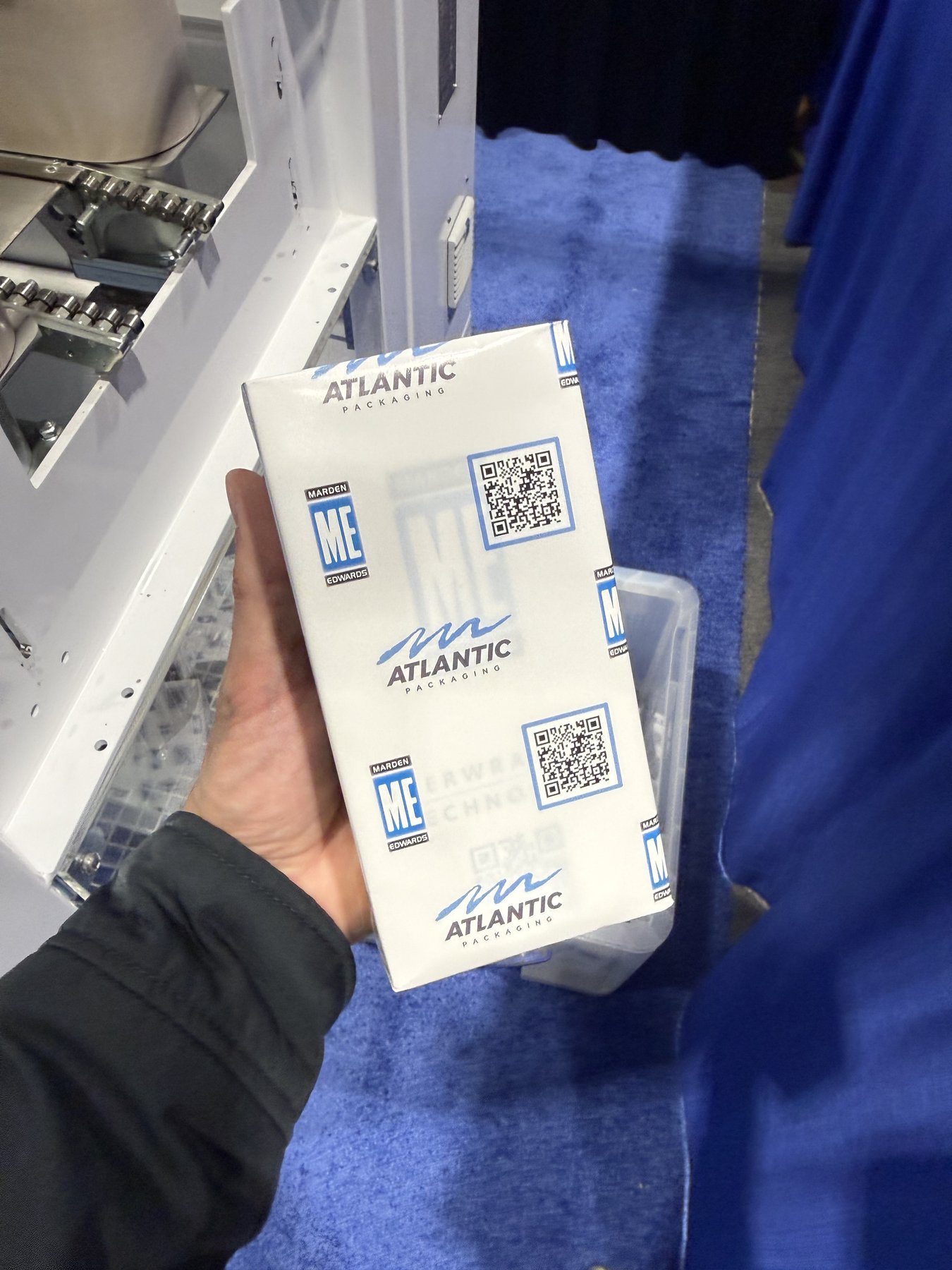A paper-overwrapped carton held up beside the machine that wrapped it